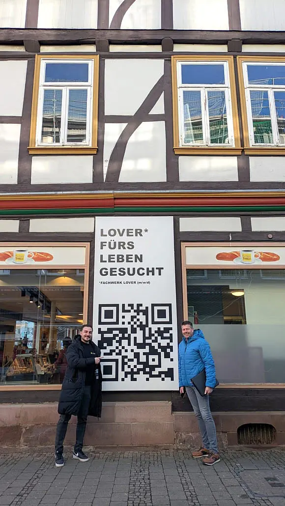 Unsere Mitarbeiter Rico und Nico vor der Tafel "Lover fürs Leben gesucht - Fachwerk Lover (m/w/d)"