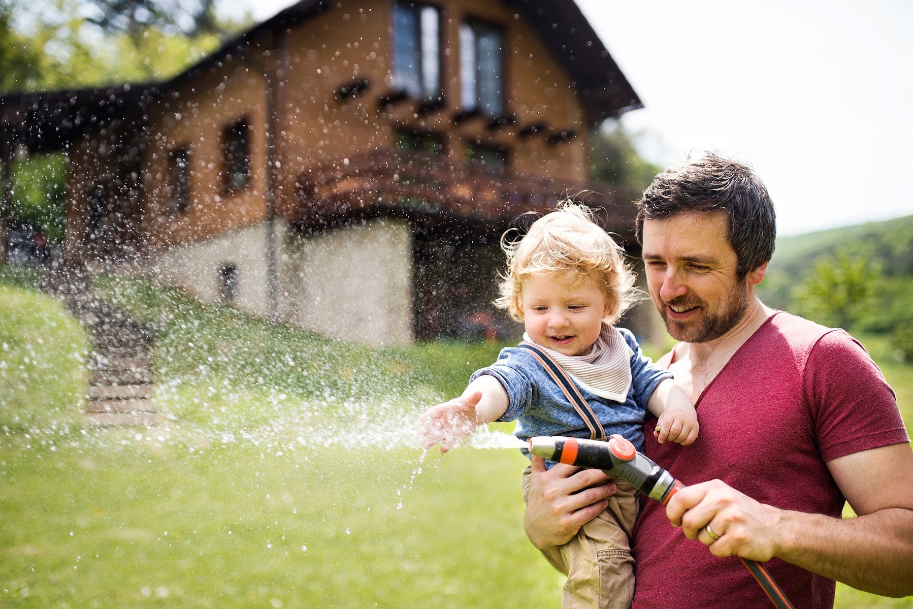 Ein Mann hat sein Baby im Arm. Sie stehen vor einem Haus auf einer Wiese und halten einen Gartenschlauch in der Hand, aus dem Wasser spritzt.