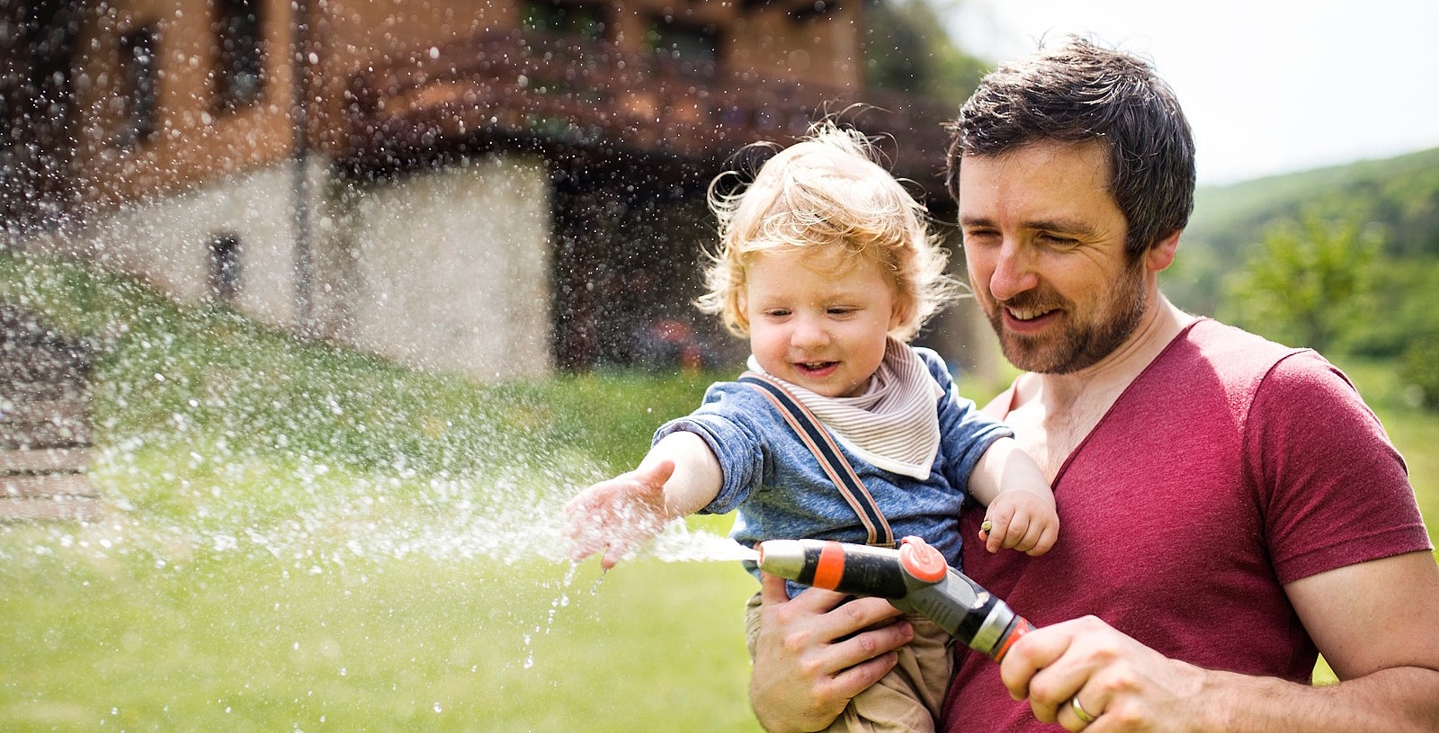Ein Mann hat sein Baby im Arm. Sie stehen vor einem Haus auf einer Wiese und halten einen Gartenschlauch in der Hand, aus dem Wasser spritzt.