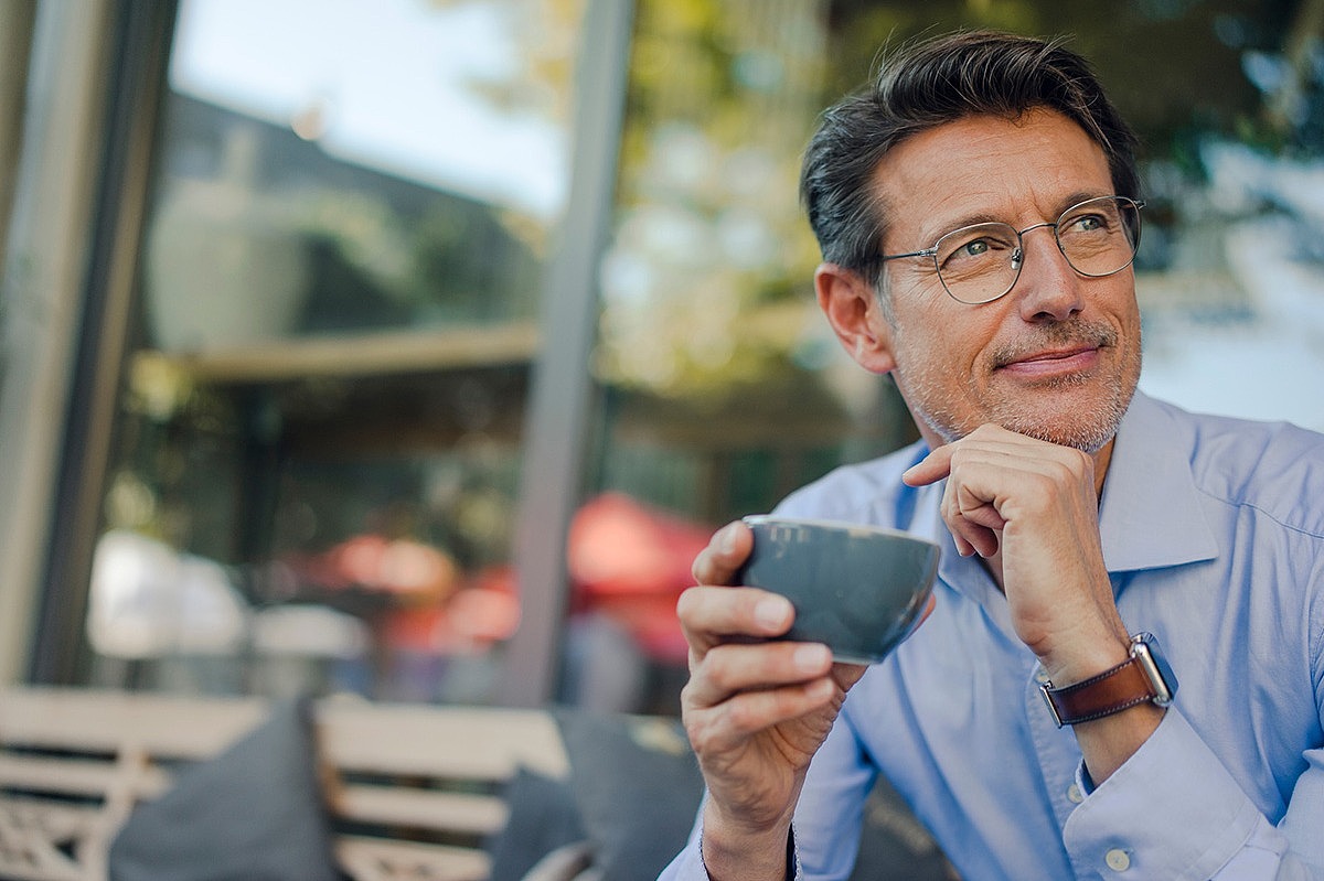 Ein Mann trinkt draußen eine Tasse Kaffee und schaut seitlich in die Kamera.