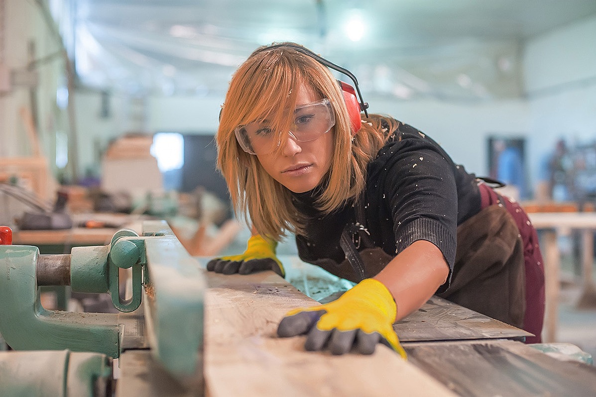 Eine junge Frau steht mit Schutzkleidung an einer Werkbank und sägt ein Stück Holz.