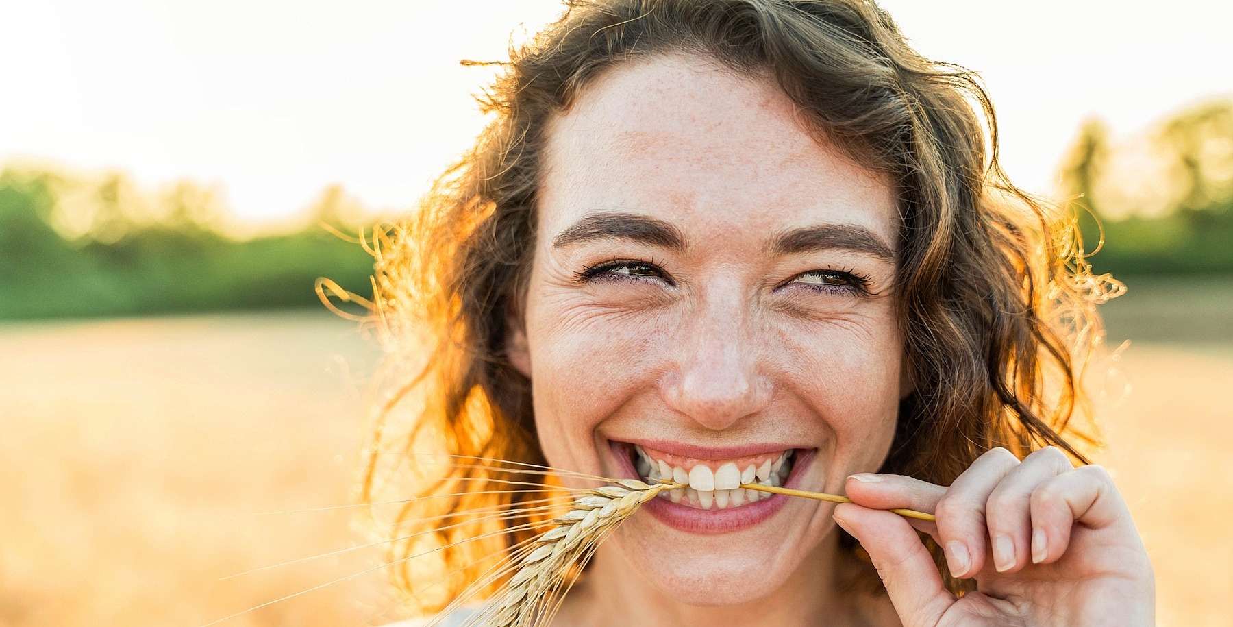 Eine junge Frau steht auf einem Feld, lächelt in die Kamera und hat eine Ähre im Mund.