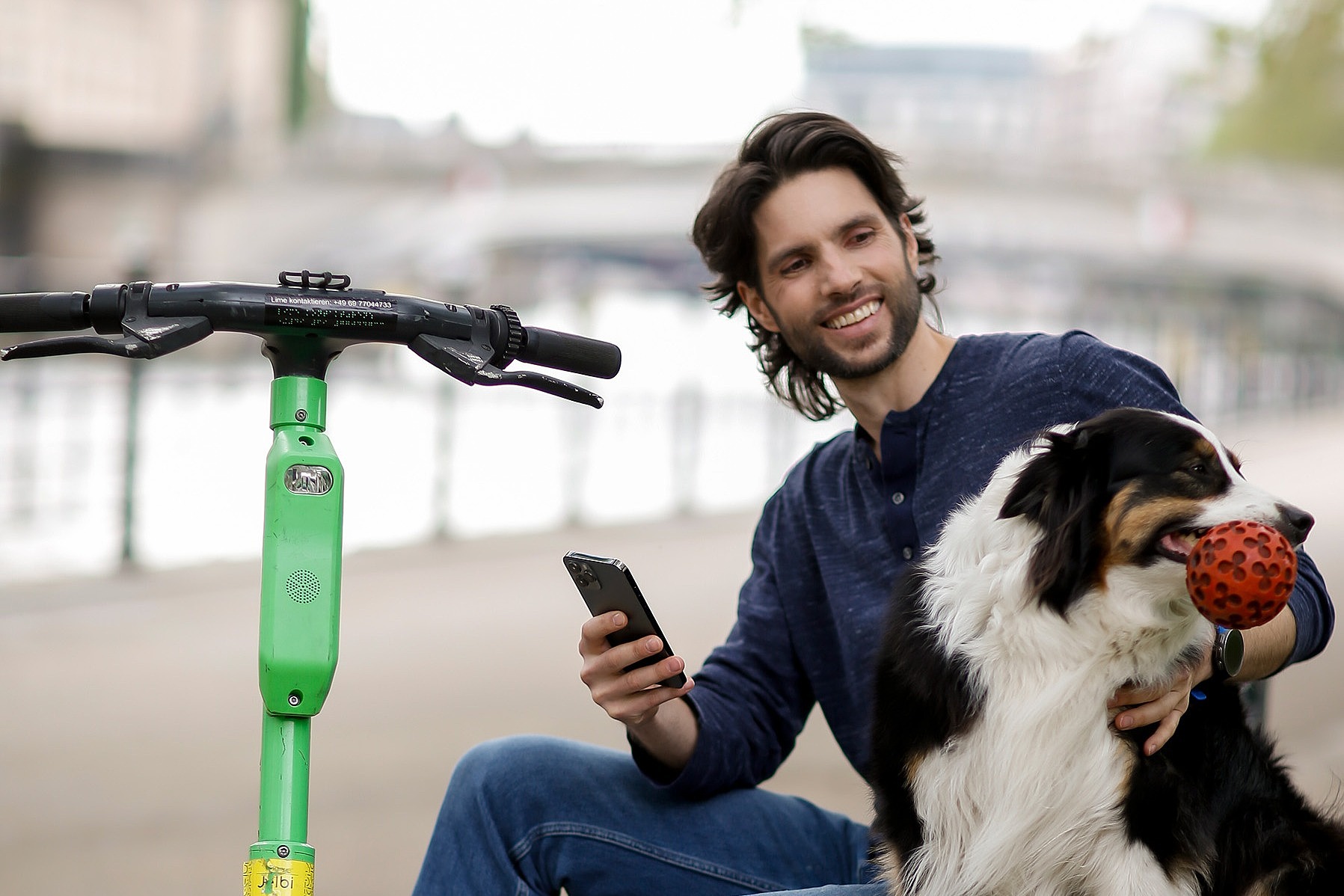 Ein junger Mann sitzt auf einer Mauer und tätschelt einen Hund. Neben ihm steht ein E-Roller.
