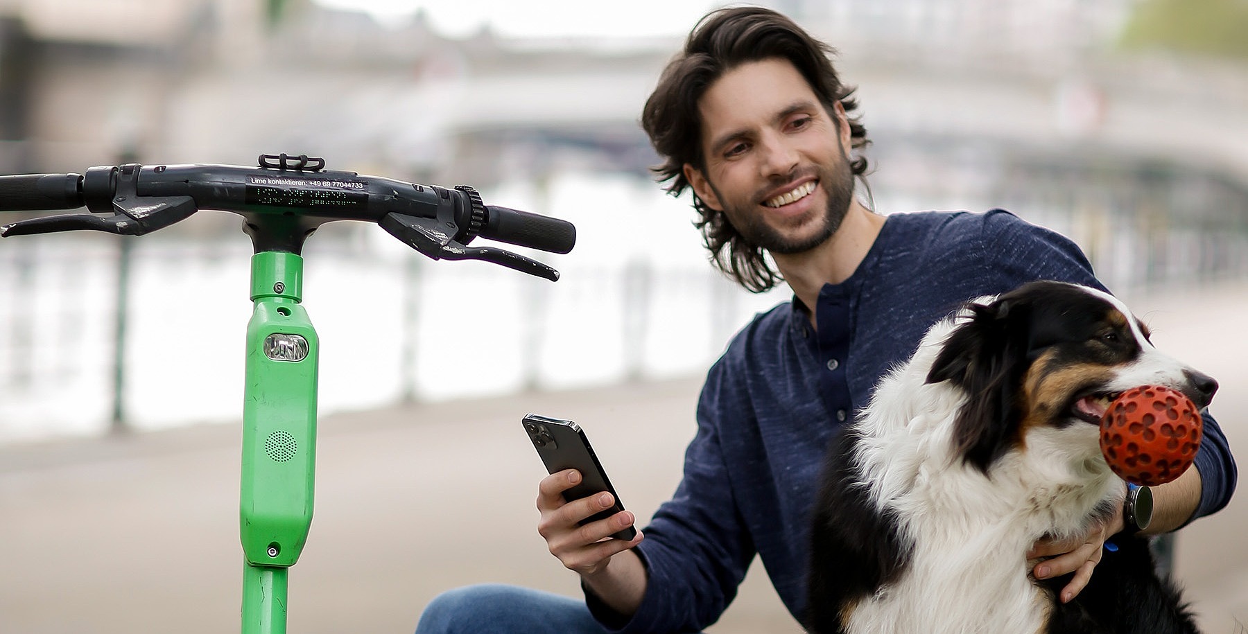 Ein junger Mann sitzt auf einer Mauer und tätschelt einen Hund. Neben ihm steht ein E-Roller.