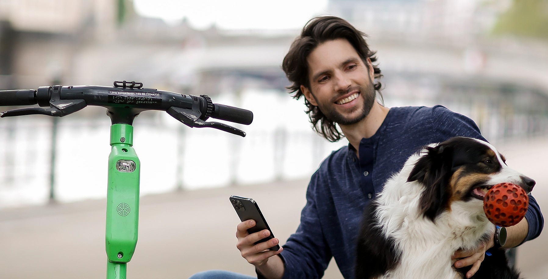 Ein junger Mann sitzt auf einer Mauer und tätschelt einen Hund. Neben ihm steht ein E-Roller.
