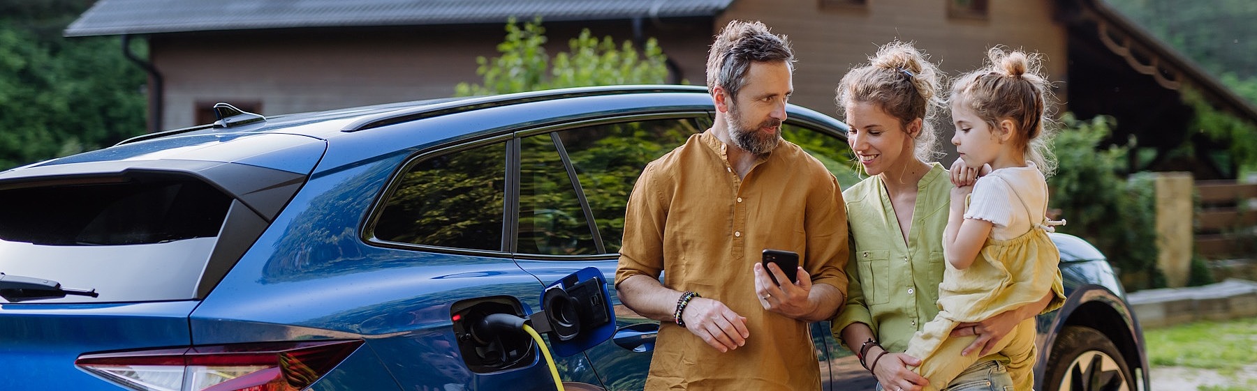 Eine junge Familie (Vater, Mutter, Kind) steht gemeinsam vor einem Auto. Im Hintergrund sieht man ein Haus.