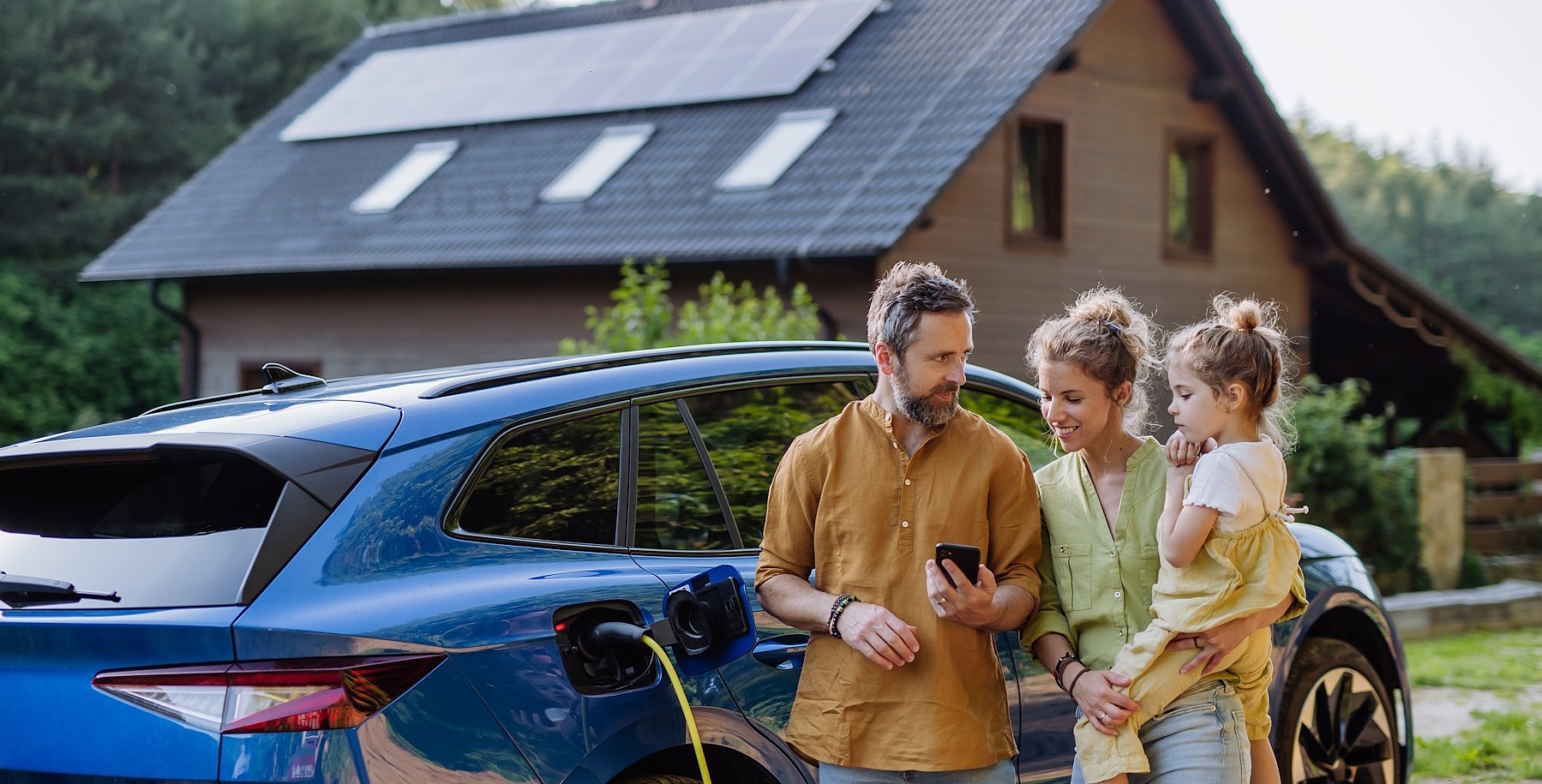 Eine junge Familie (Vater, Mutter, Kind) steht gemeinsam vor einem Auto. Im Hintergrund sieht man ein Haus.