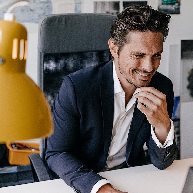 Ein Mann sitzt in seinem Büro am Schreitisch und schaut lächelnd auf den Bildschirm seines Computers.