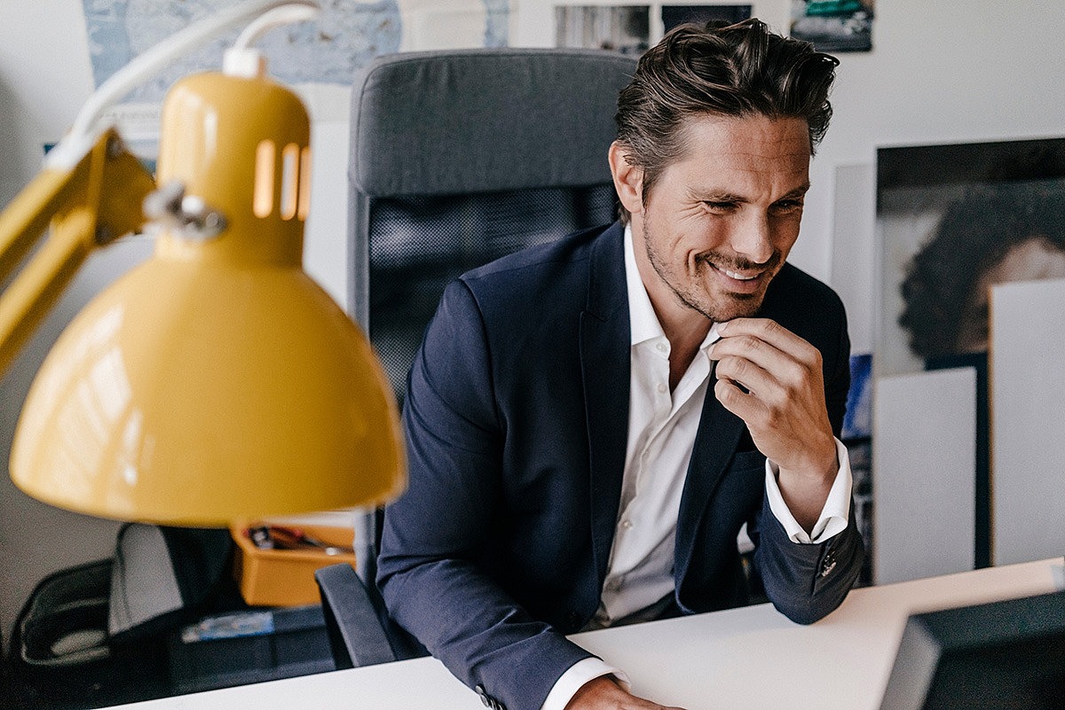 Ein Mann sitzt in seinem Büro am Schreitisch und schaut lächelnd auf den Bildschirm seines Computers.