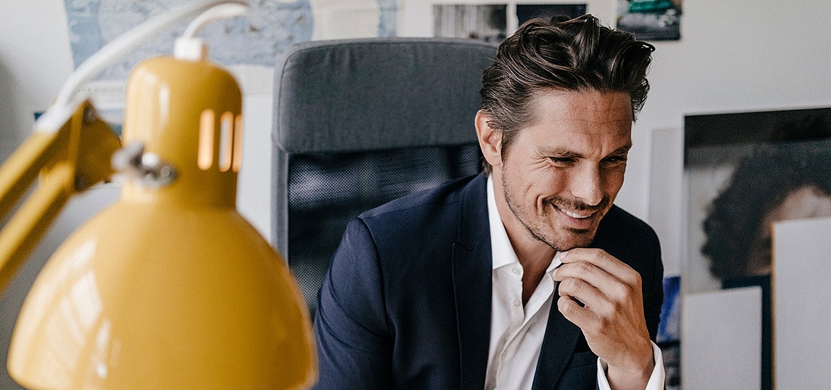 Ein Mann sitzt in seinem Büro am Schreitisch und schaut lächelnd auf den Bildschirm seines Computers.