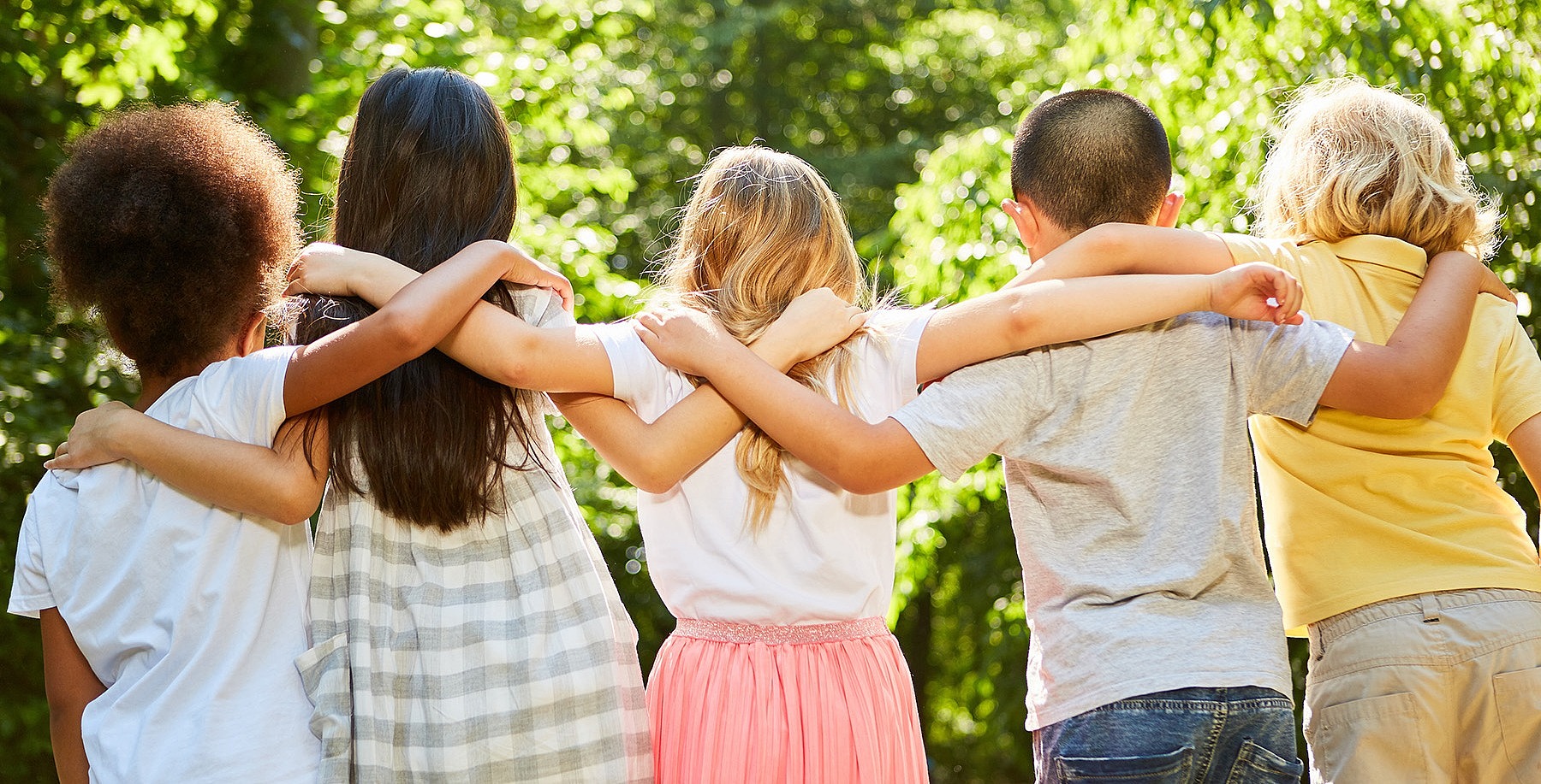 Gruppe von fünf Kindern steht mit dem Rücken zur Kamera. Sie haben sich gegenseitig die Arme um die Schultern gelegt.