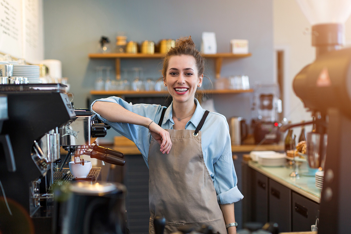 Eine junge steht in einem Cafe hinter der Theke. Sie hat eine Schürze um und legt den Arm lässig auf eine Kaffeemaschine.