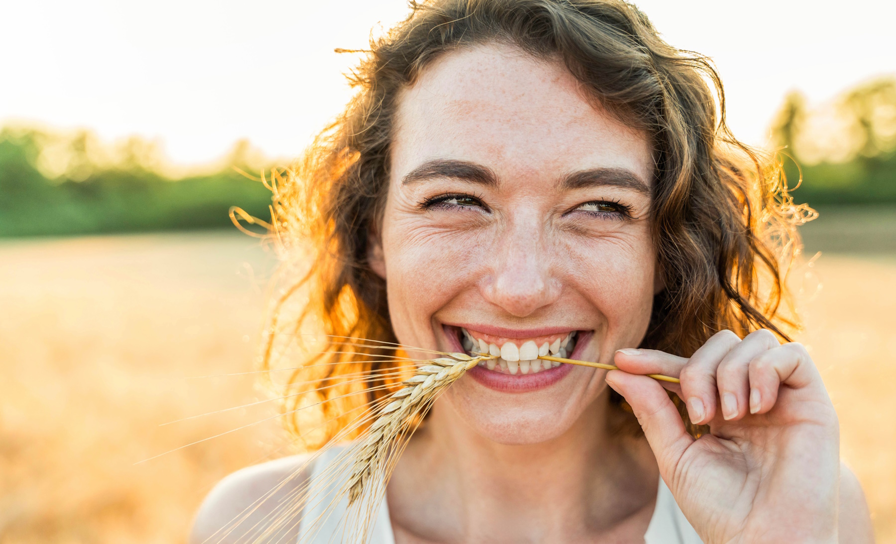 Eine junge Frau steht auf einem Feld, lächelt in die Kamera und hat eine Ähre im Mund.